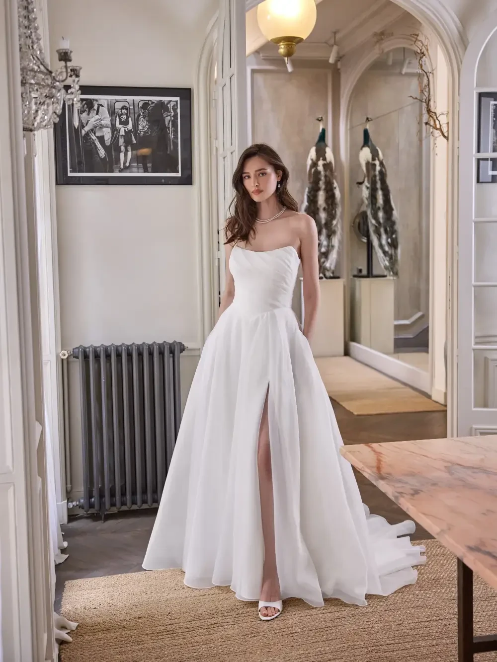 A woman in a strapless white wedding dress with a thigh-high slit stands in a stylish interior, featuring elegant decor and a vintage photograph on the wall.