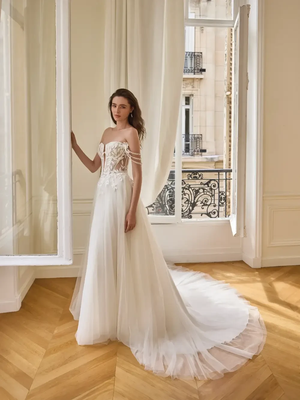A model wearing a strapless wedding dress with a long train, standing by a large window in a bright, airy room.