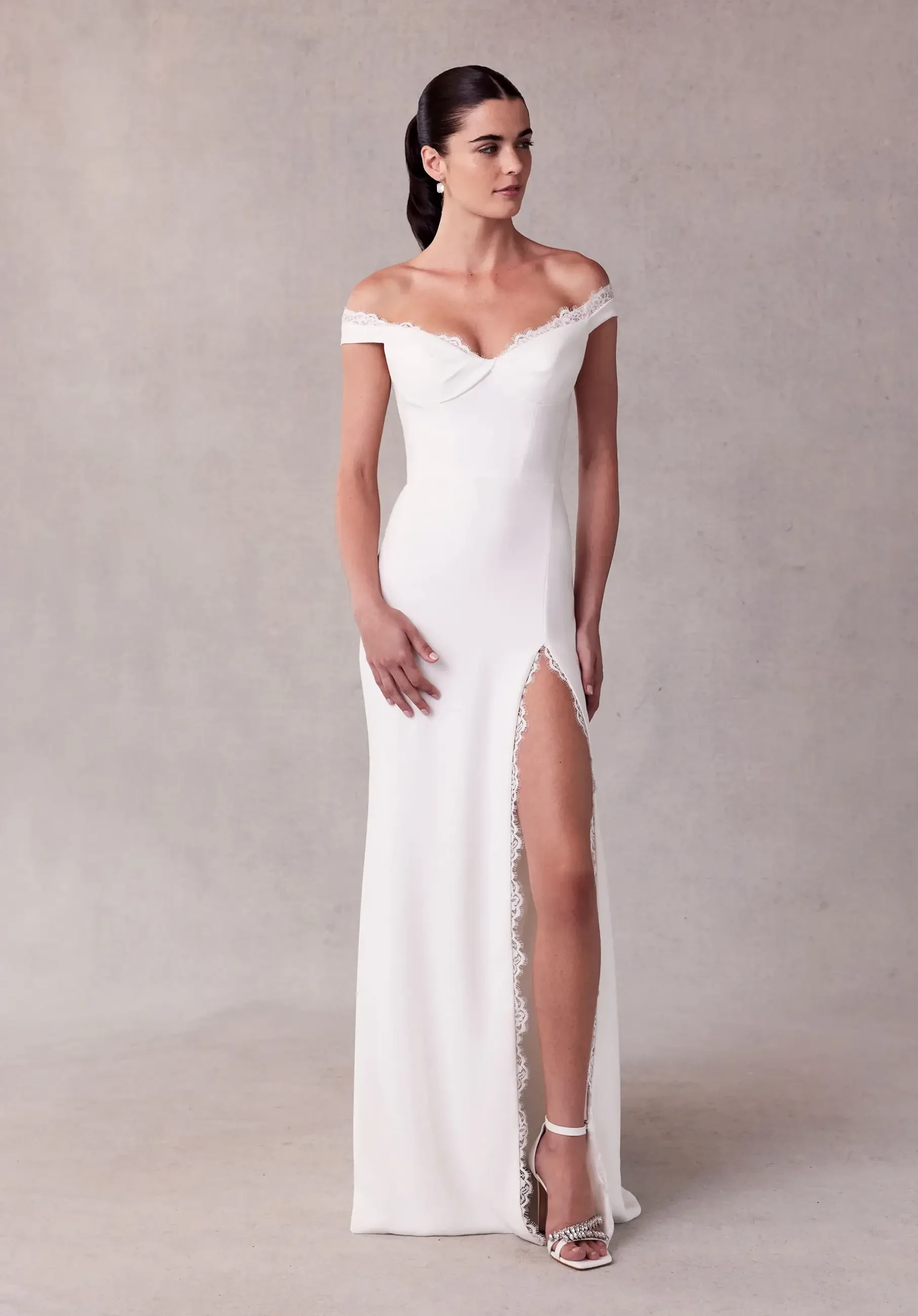 A woman in an elegant off-the-shoulder white gown with lace trim and a high leg slit stands against a minimalist gray backdrop. She exudes sophistication.