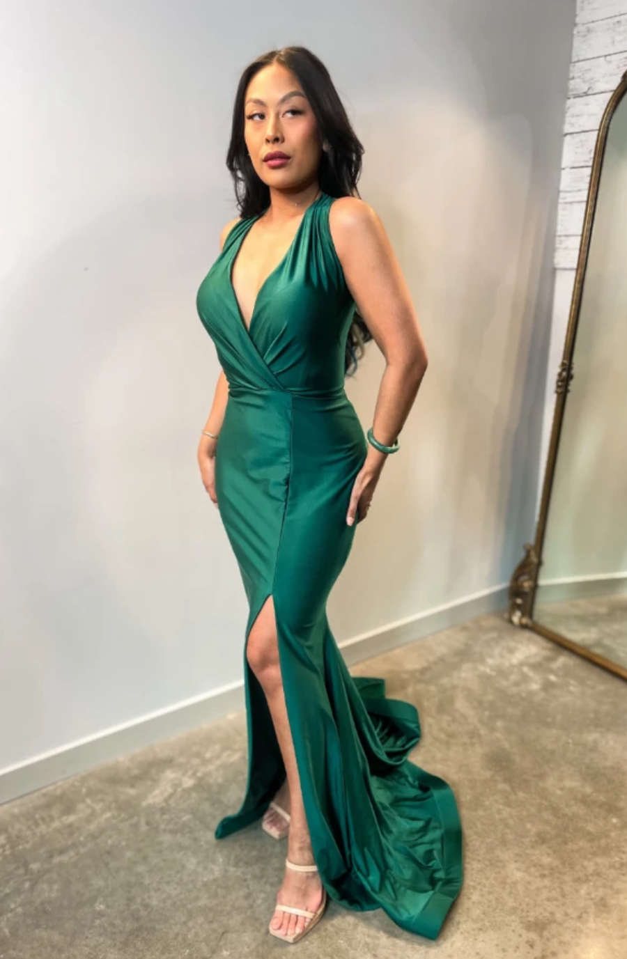 Woman in an elegant green dress with a deep V-neckline and high slit, posing confidently near a mirror on a concrete floor, exuding sophistication.