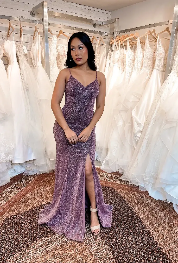 A woman in a sparkly purple dress with a slit stands in front of a backdrop of wedding dresses.