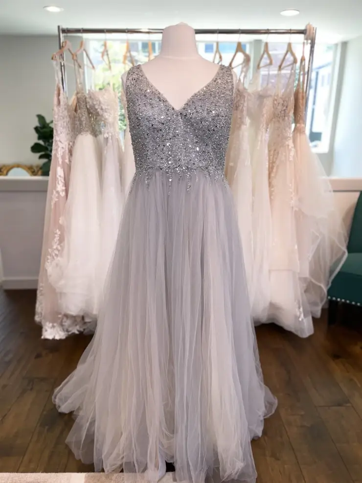 A sparkling silver and gray gown with a fitted bodice and a flowing tulle skirt, displayed on a mannequin in front of several other dresses.