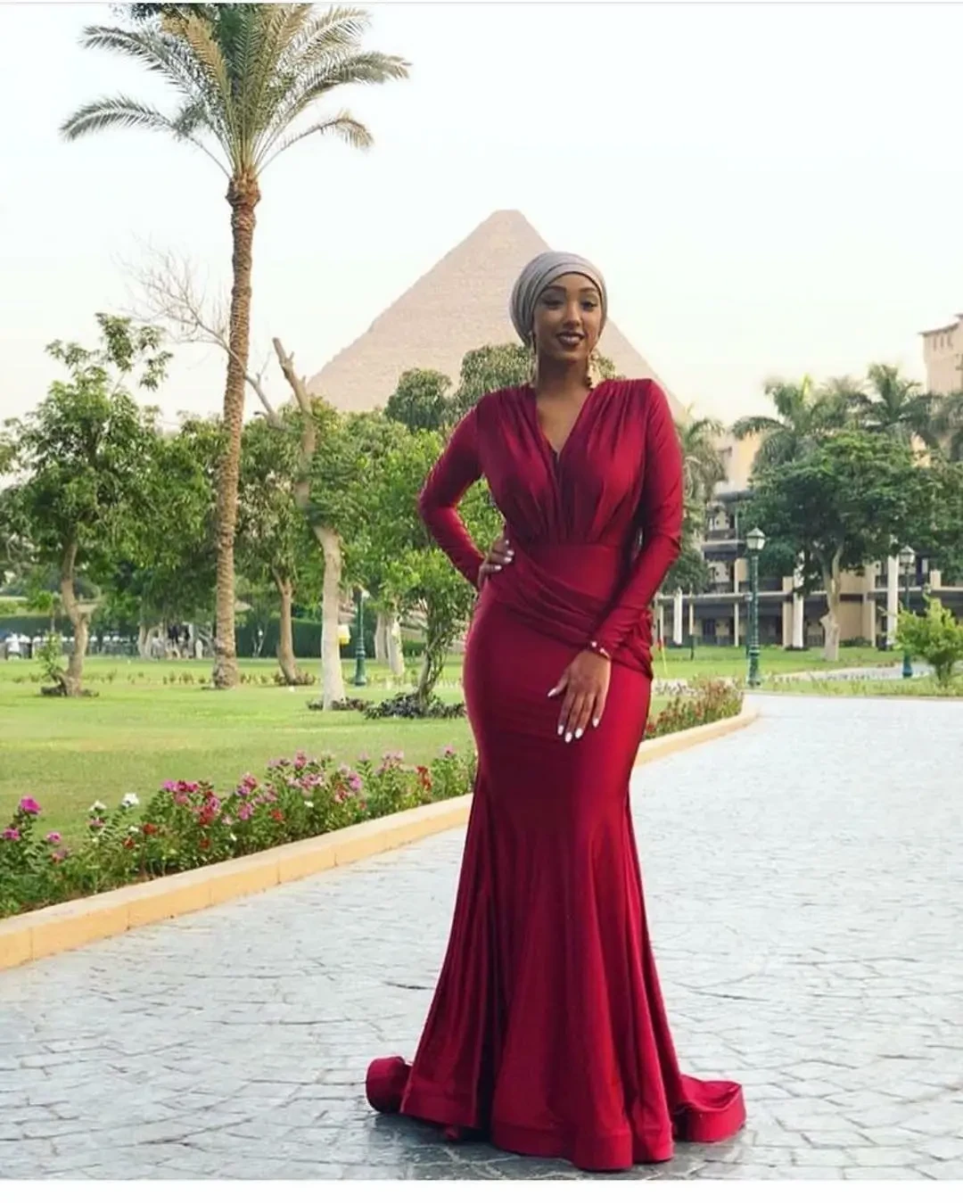 A woman in a red long-sleeved gown stands outdoors with palm trees and the pyramids in the background.