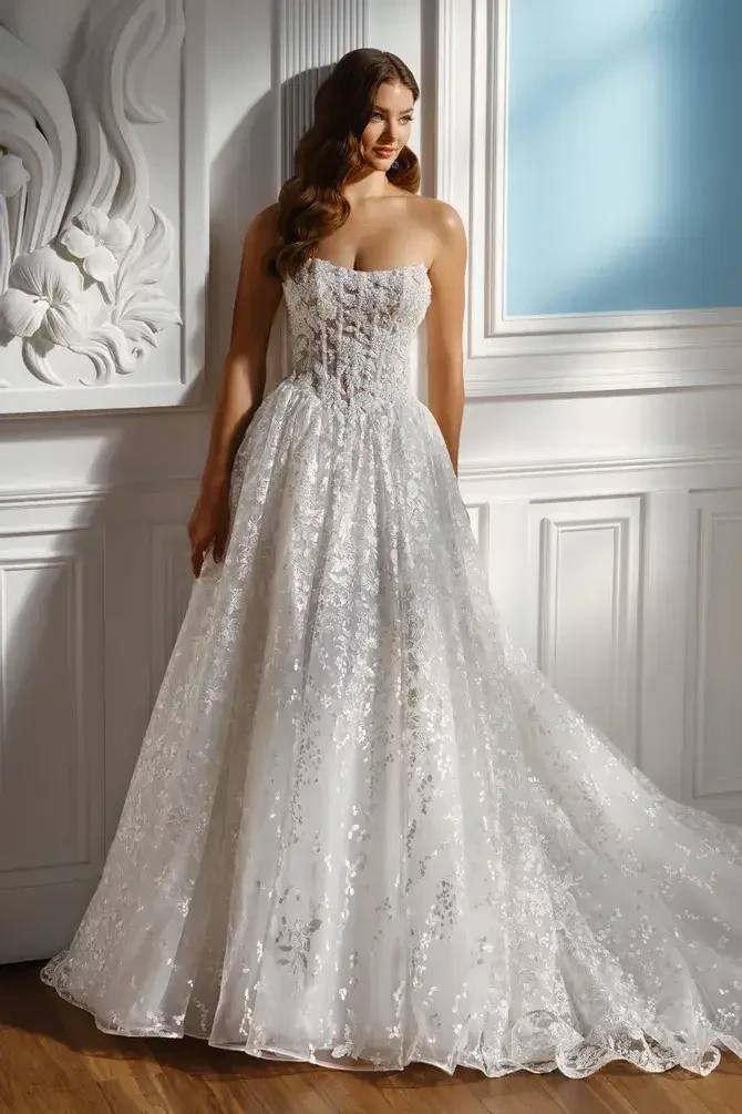 A woman wearing a strapless, floor-length white wedding gown with intricate lace detailing, standing against an elegantly designed wall.