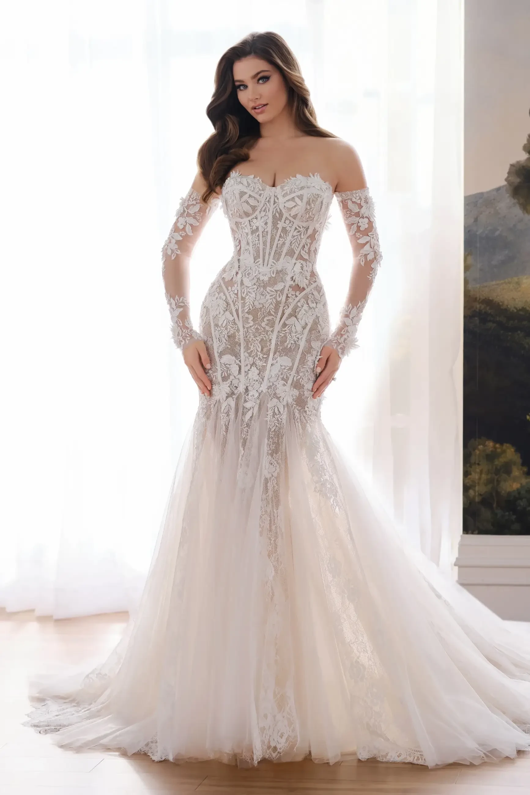 A woman wearing an intricate off-the-shoulder lace wedding gown with sheer sleeves stands in front of a softly lit window, exuding elegance and grace.
