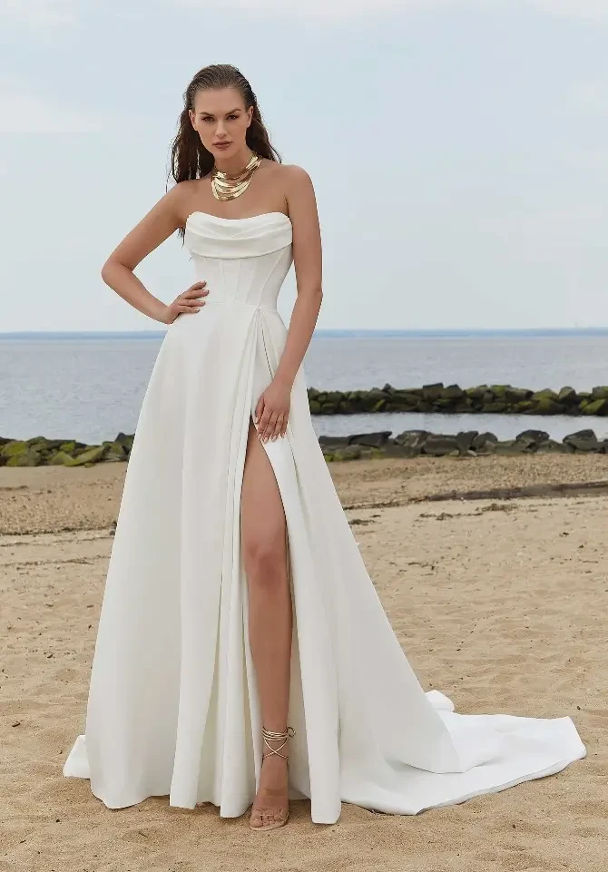 A model poses in a strapless white gown with a high slit, standing on a sandy beach beside the water.
