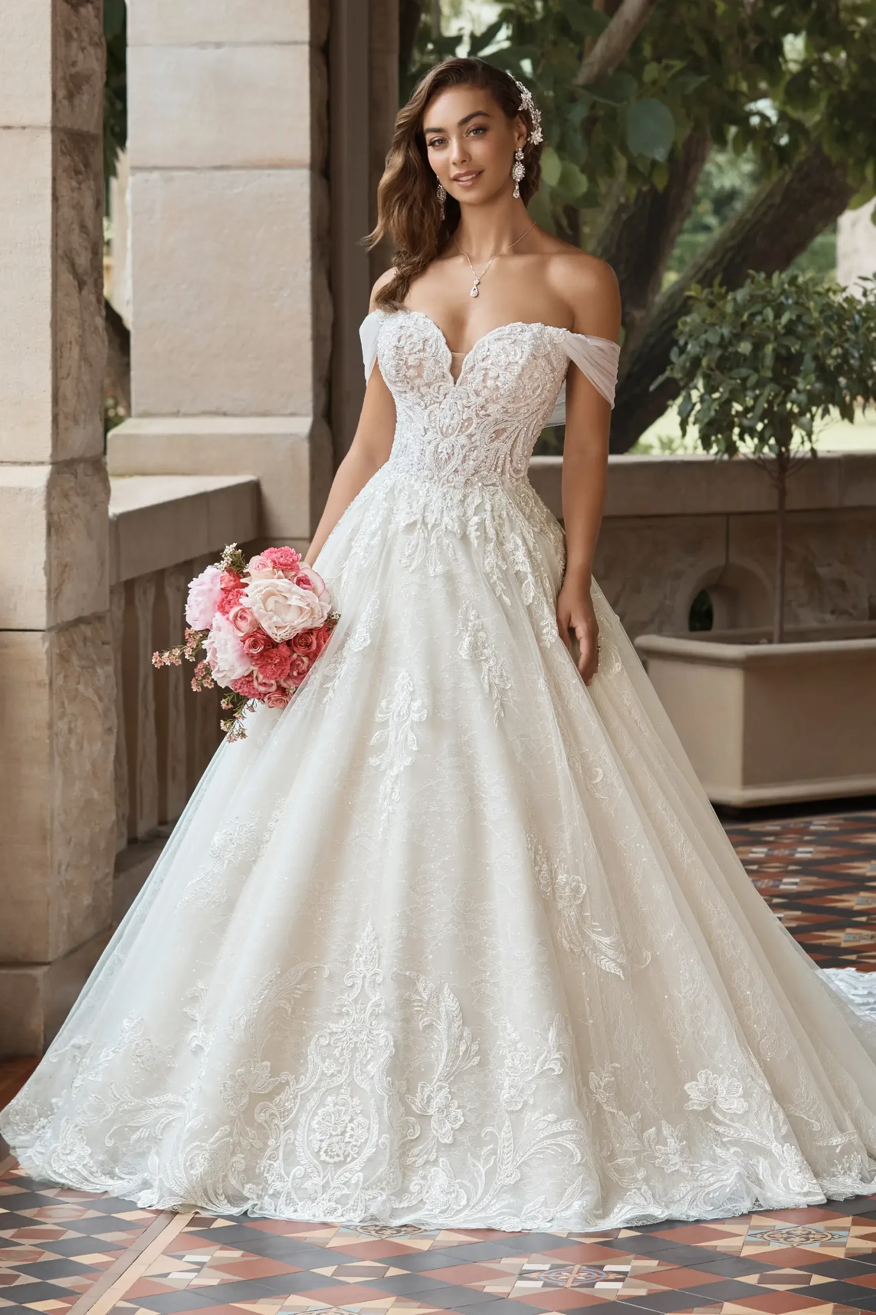 A bride in a lace off-the-shoulder wedding gown stands on a tiled floor, holding pink and white flowers, with greenery in the background. Elegant and romantic.