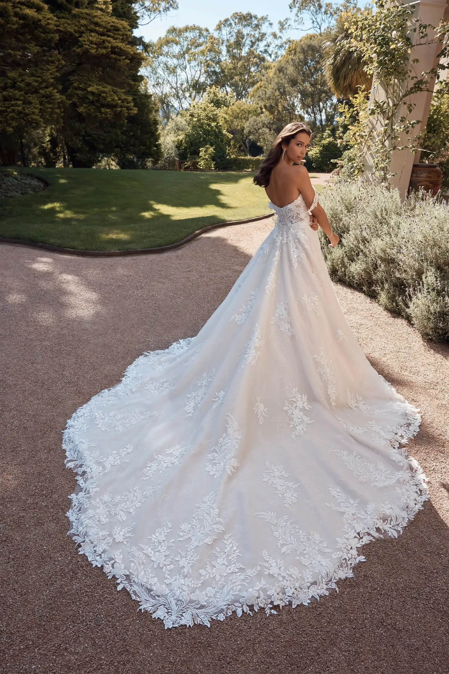 Model is wearing a regal off-the-shoulder ballgown with a sweetheart neckline, low lace-trimmed back, and a full chapel-length train made of misty tulle and organza.