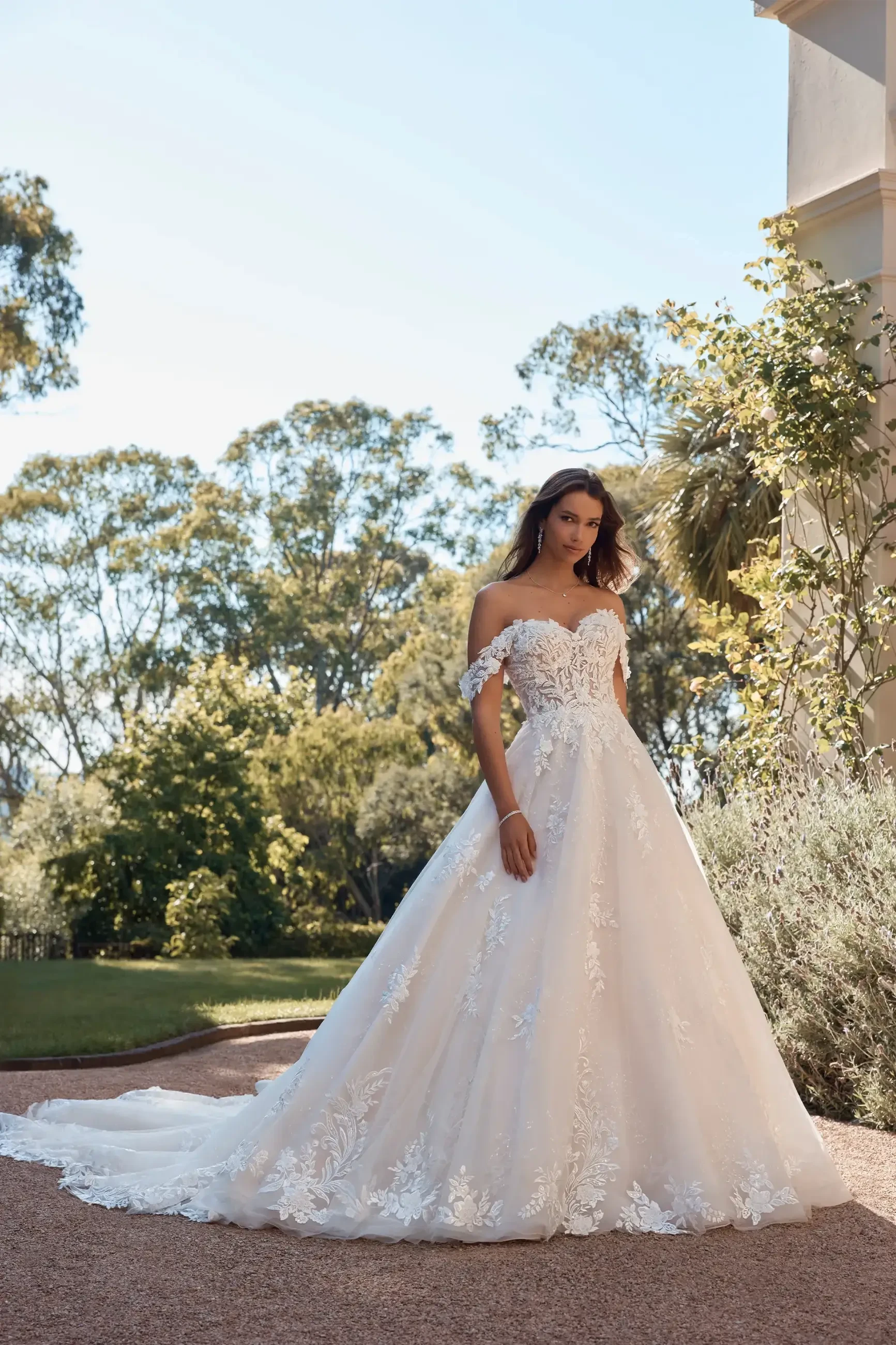 Model is wearing a regal off-the-shoulder ballgown with a sweetheart neckline, low lace-trimmed back, and a full chapel-length train made of misty tulle and organza.