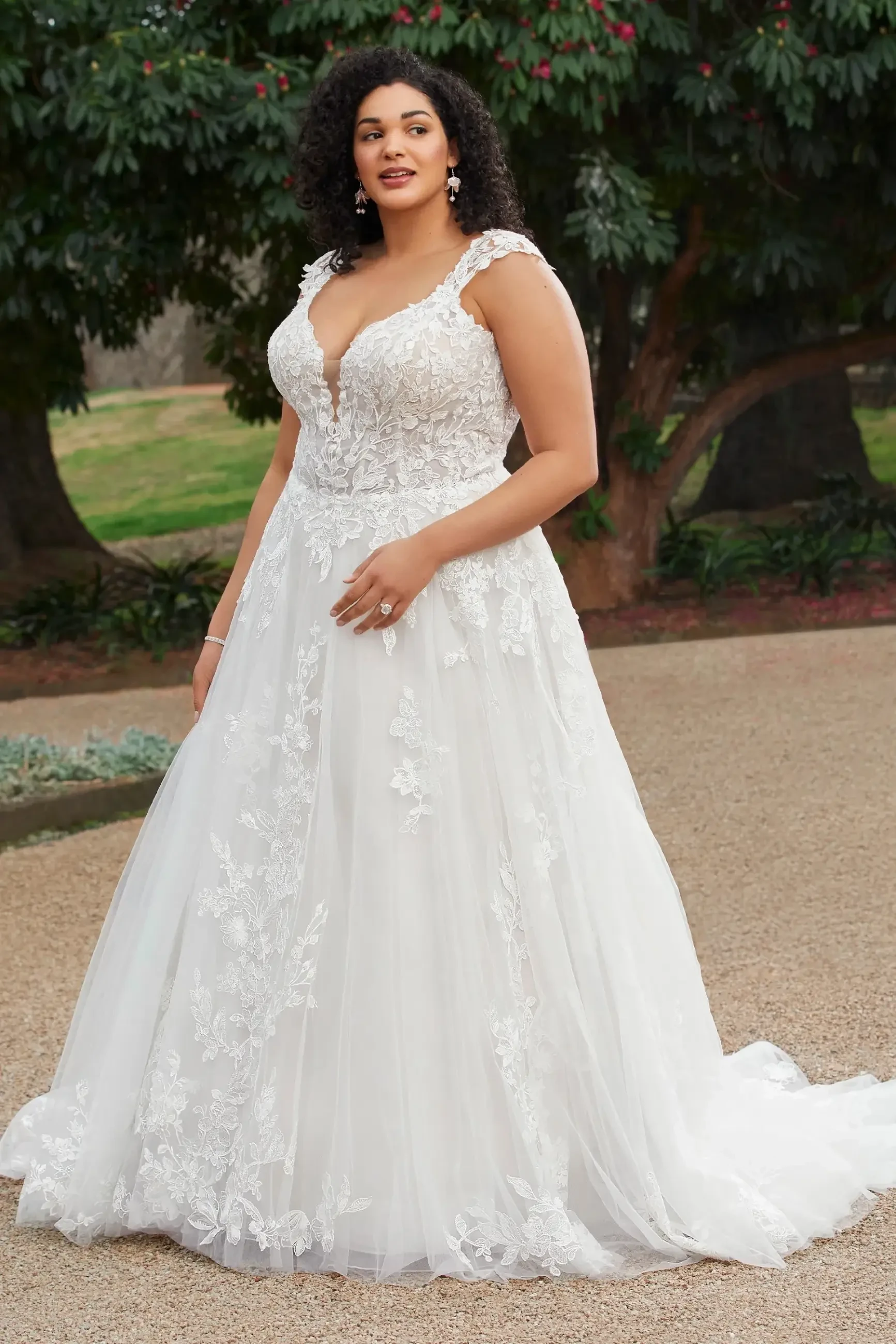 A woman stands outdoors in an elegant white wedding dress with lace detailing, exuding confidence. She is surrounded by greenery and trees.