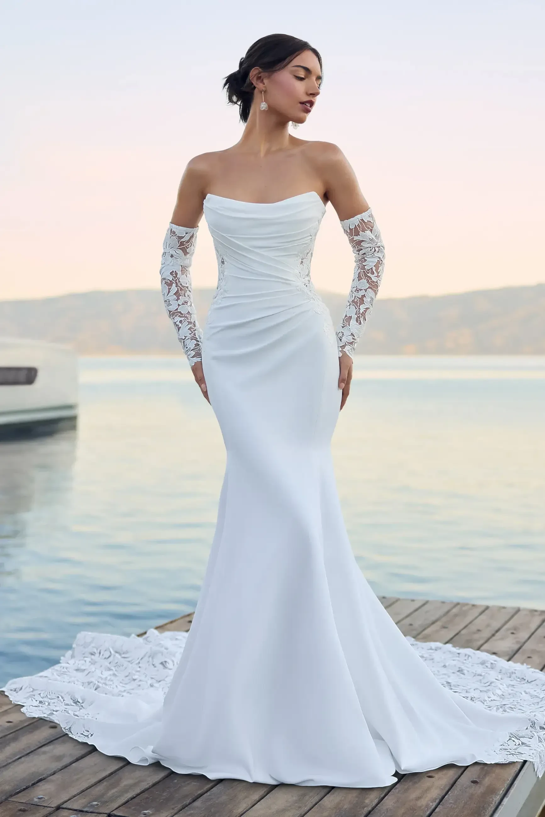 Model is wearing a romantic crepe wedding dress with an illusion lace back, off-the-shoulder long sleeves, draped bodice, and a dramatic laser-cut train.