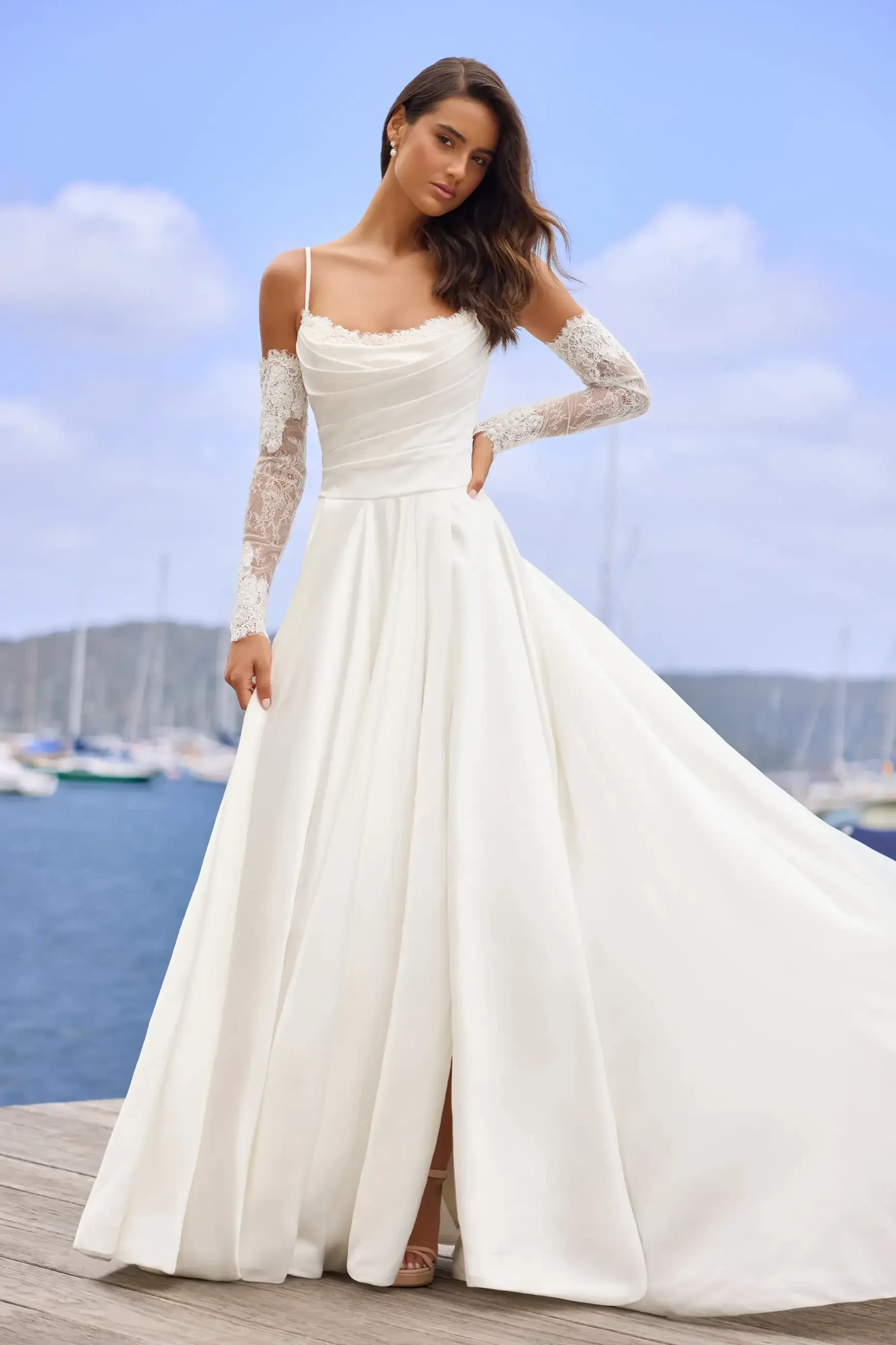 A woman in a flowing white wedding dress with lace sleeves stands on a boardwalk by the sea. The background features a blue sky and sailboats.