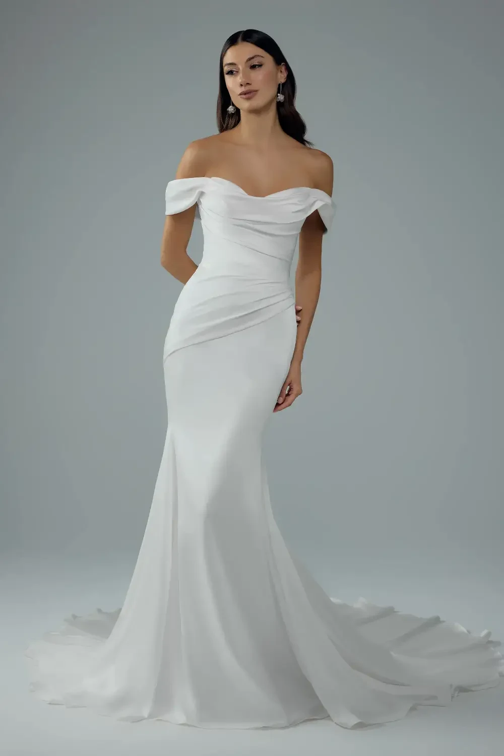 A woman wearing an elegant, off-the-shoulder white gown with a fitted silhouette and a flowing train, standing gracefully against a neutral background.