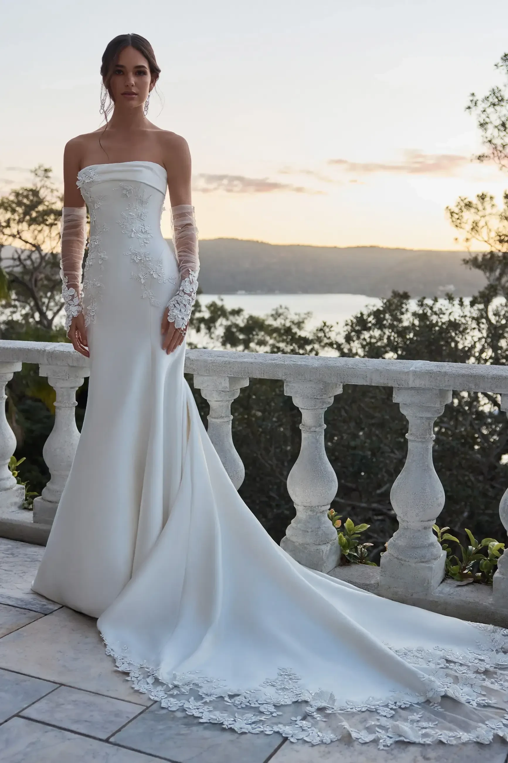 A woman in a strapless, elegant white wedding gown with lace details stands on a terrace at sunset, evoking romance and serenity.