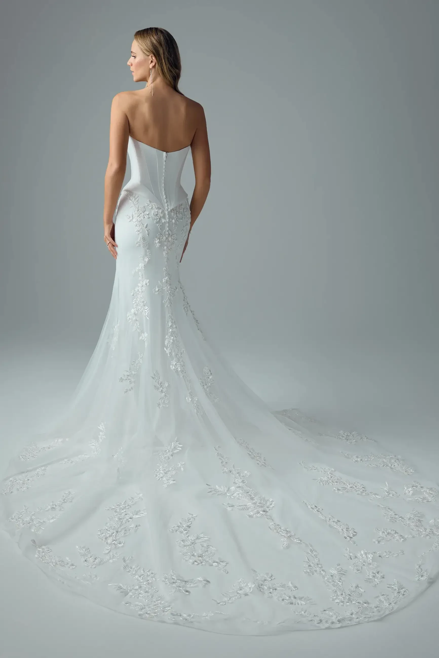 A woman in a strapless, white lace bridal gown with delicate floral embroidery stands facing away, showcasing the elegant train in a serene setting.