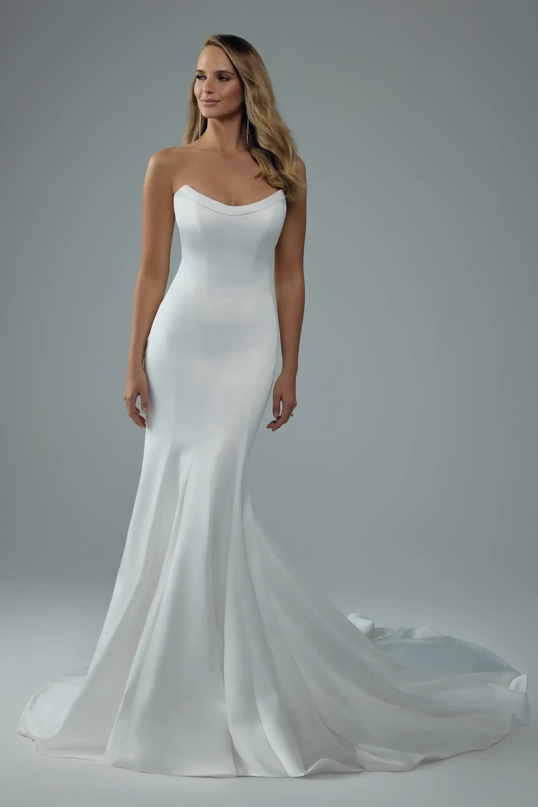 A woman in a strapless, form-fitting white wedding gown with a subtle train stands confidently. The background is a soft gray, creating an elegant tone.