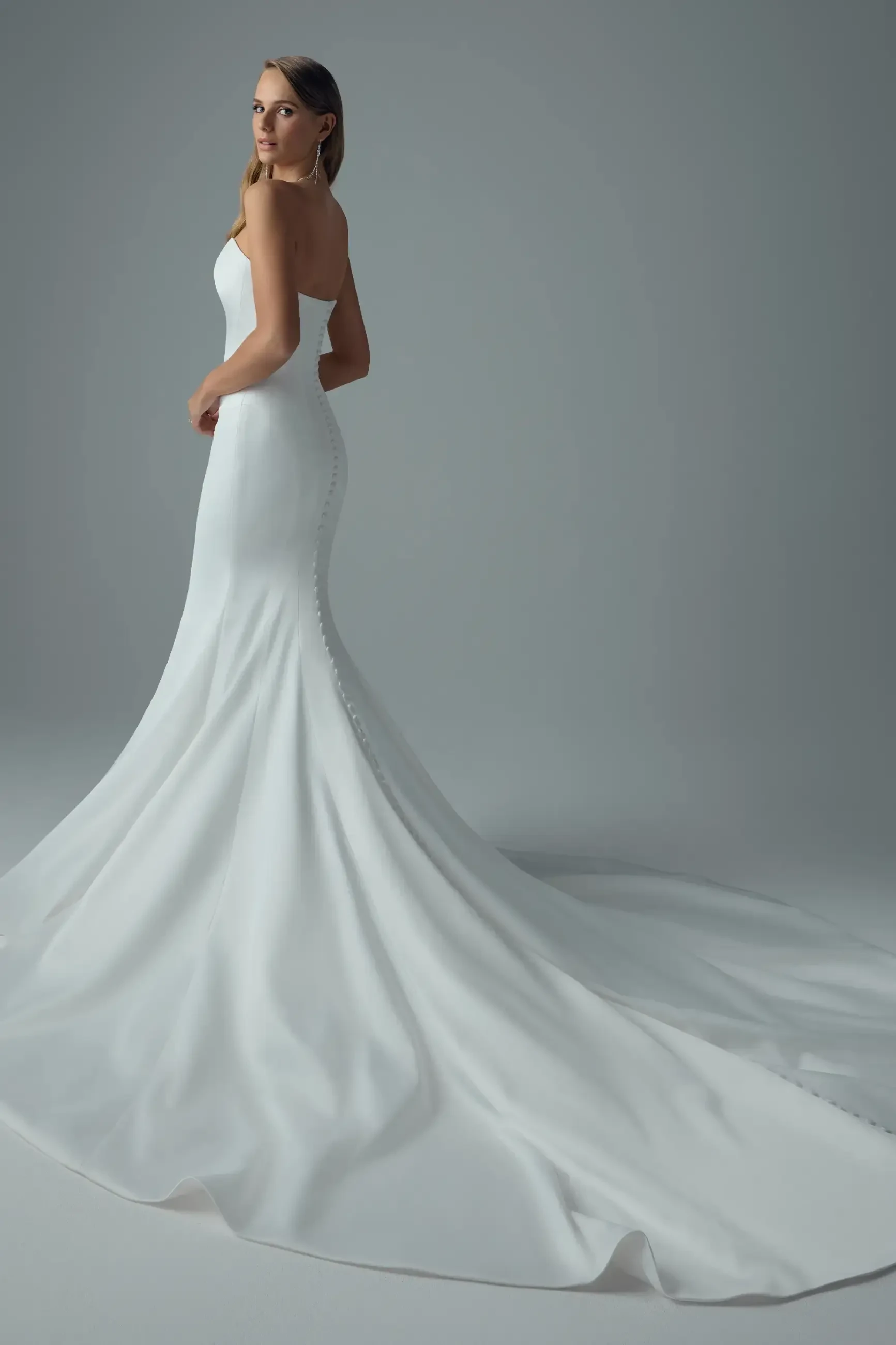 A woman in a sleek, strapless white wedding gown with a long train stands in profile. She gazes back, exuding elegance and tranquility in a soft gray setting.