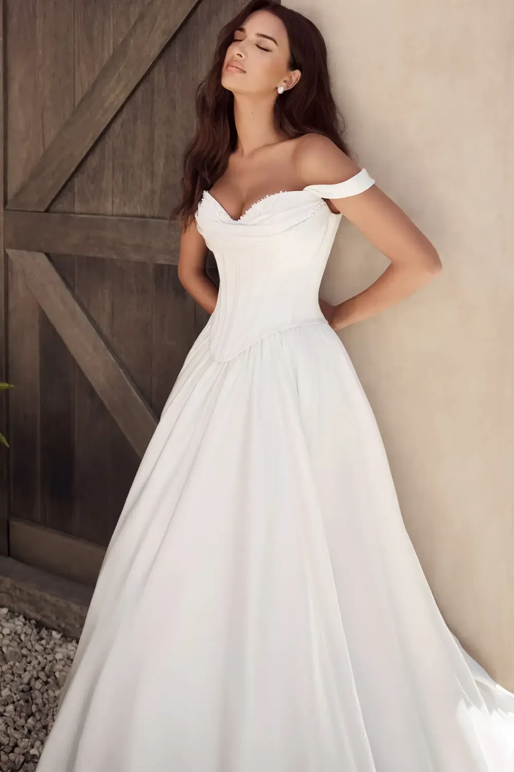 A woman wearing an elegant off-the-shoulder white wedding gown, standing gracefully against a wooden backdrop.